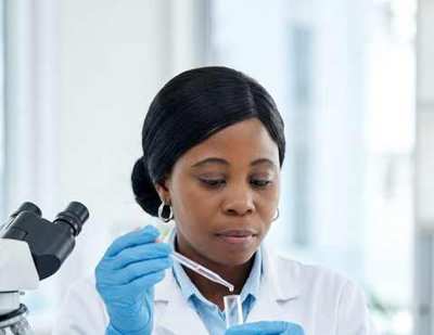 A laboratory scientist in a white coat and blue gloves holding a test tube beside a microscope
