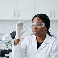 A laboratory scientist in a white coat, safety glasses and gloves holding a sample tube beside a microscope